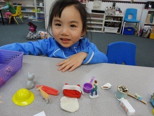 Young girl smiles at the camera, surrounded by toys on a table in a classroom