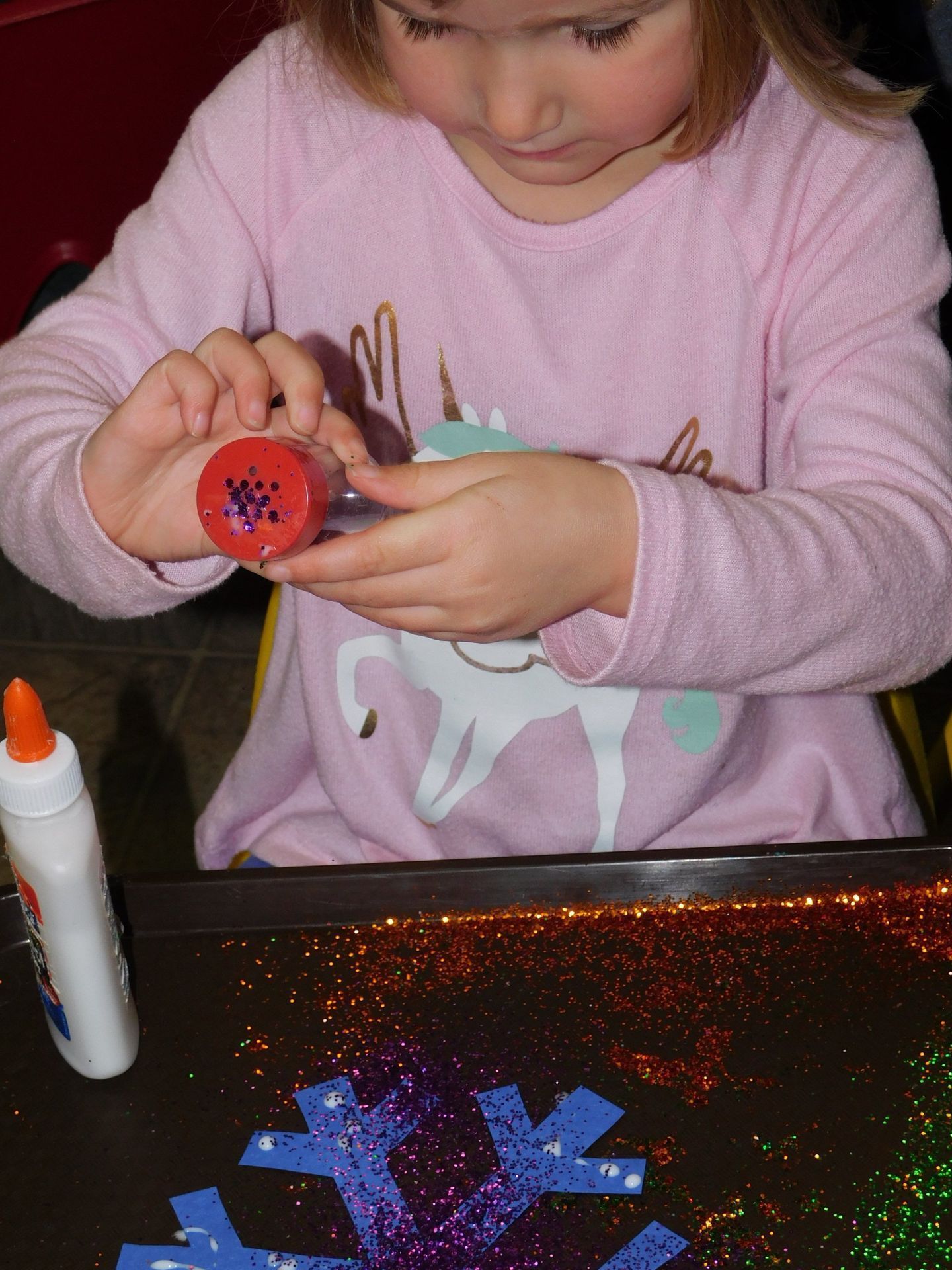 Girl in pink shirt decorating a red cylinder with glitter, seated at a craft table with glue and glitter