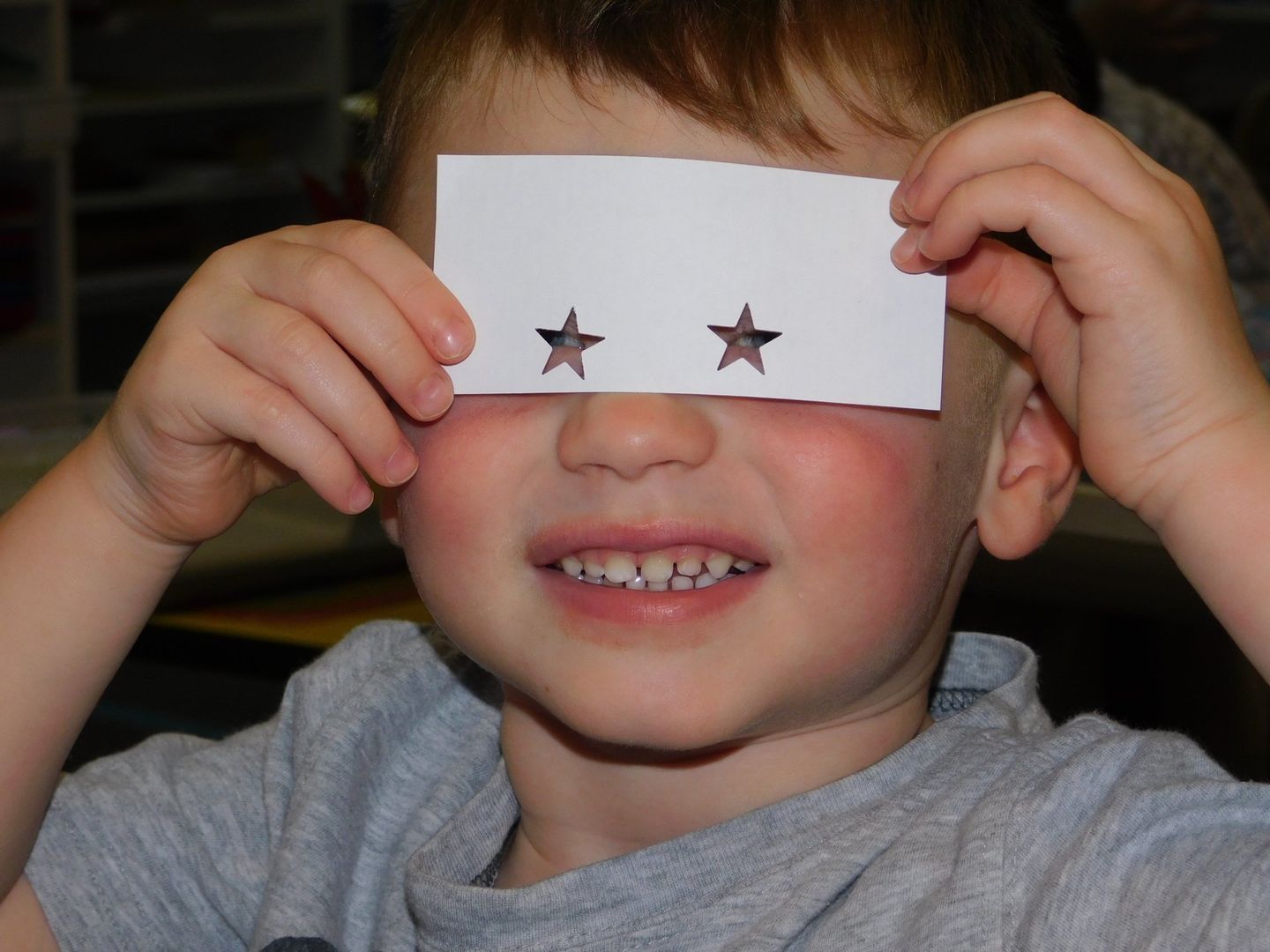 Young child smiling, holding a white paper with star-shaped holes over their eyes