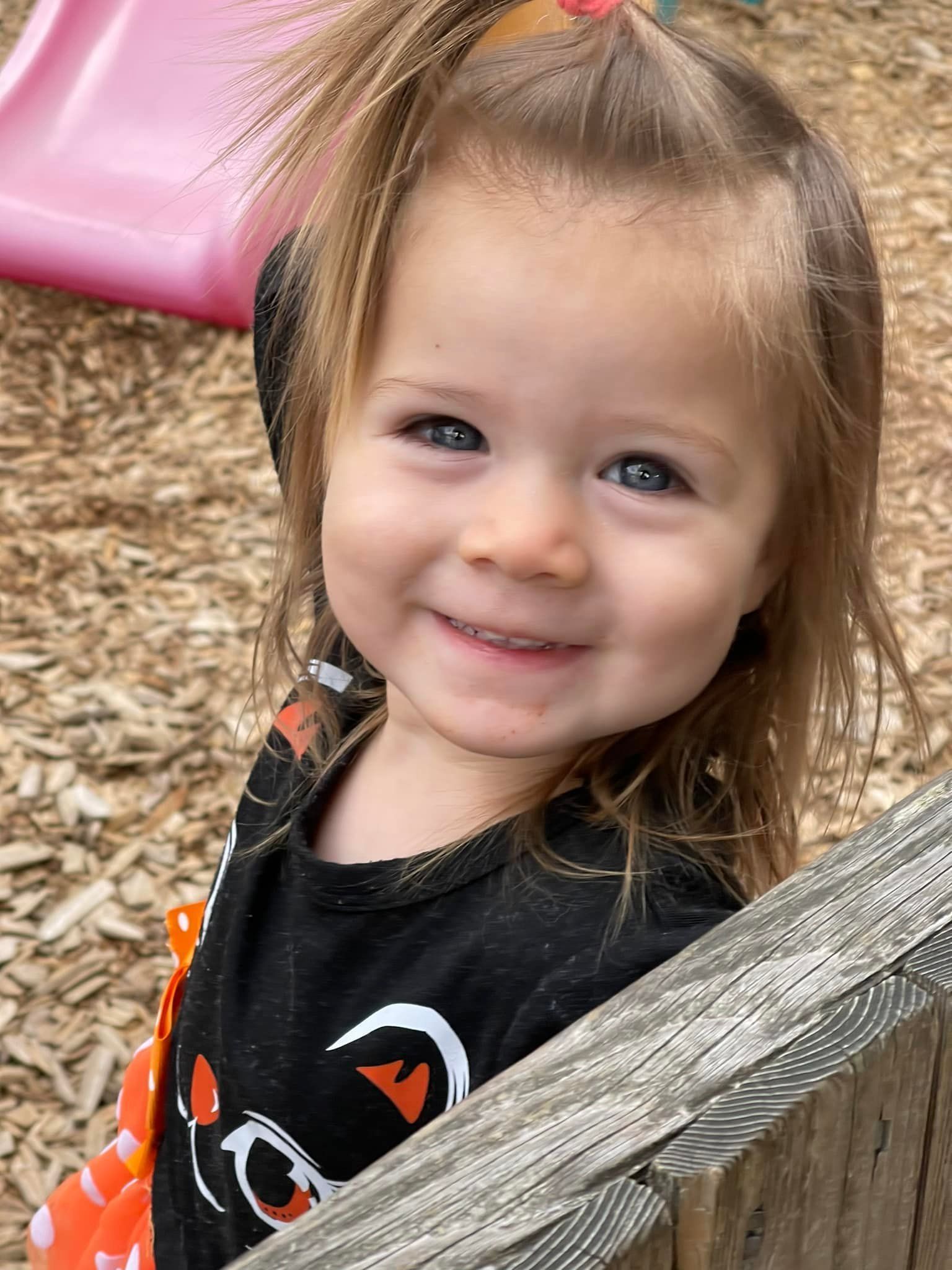 Girl with blue eyes smiles, wearing black cat shirt, standing near a wooden fence, outdoors