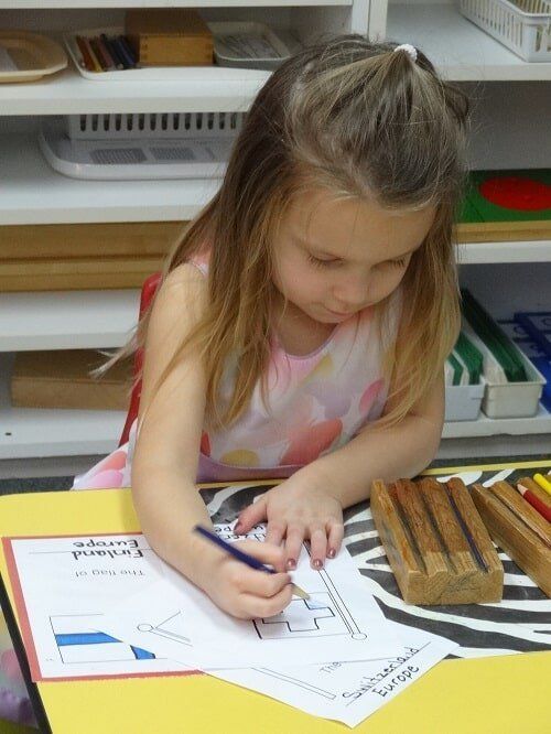 A young girl colors a worksheet about Europe at a desk, focused and concentrated
