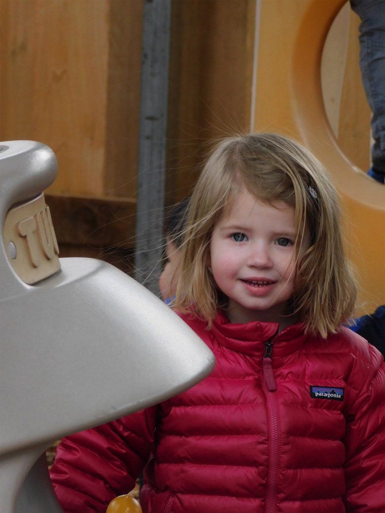 Young girl with blonde hair smiles in a red puffer jacket at a playground.