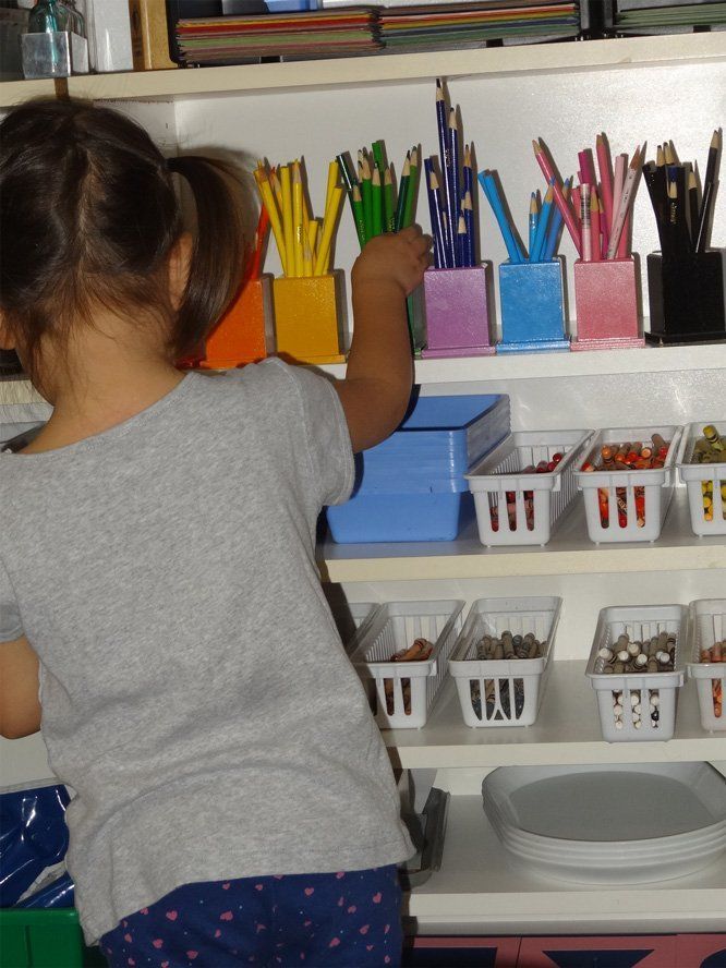 A young child reaching for colored pencils in a rainbow-colored container on a shelf