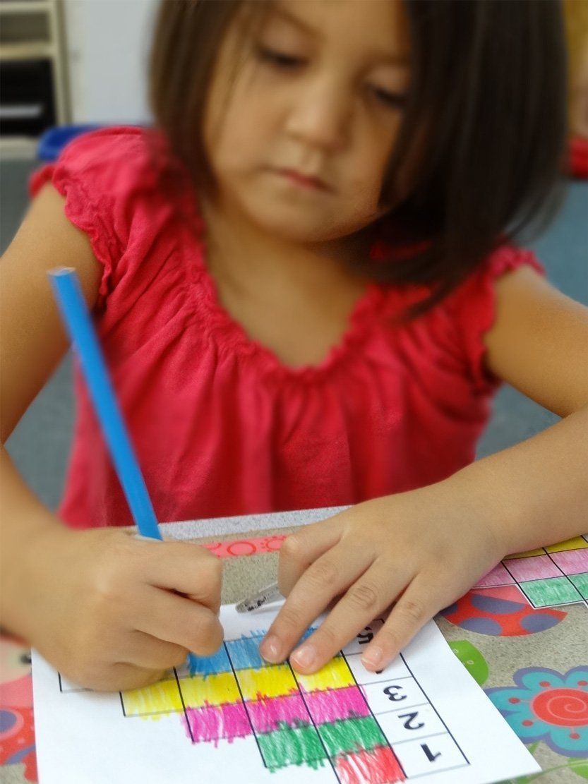 Girl in pink top coloring a grid with a blue pencil at a table