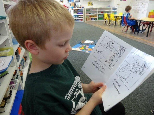 A young boy with blond hair reads a book in a classroom, focused expression