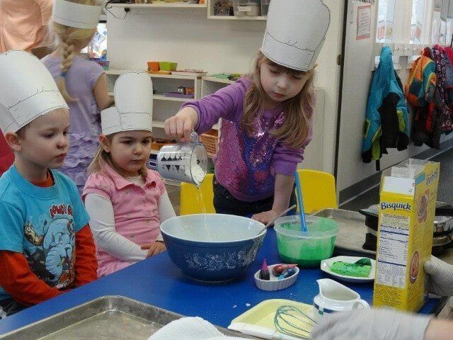 Children in chef hats pouring batter in a classroom, preparing food