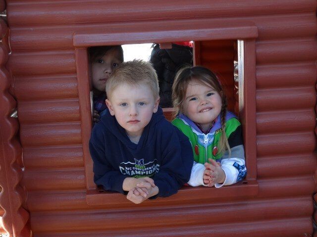 Three children, two boys and a girl, peering out of a wooden playhouse window, smiling