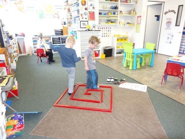 Two young boys playing inside a classroom, standing within a red square on a brown mat