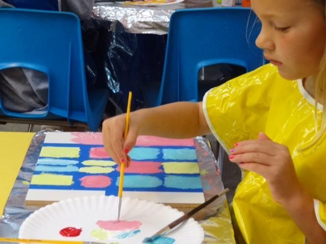 Young girl in yellow smock paints colorful rectangles on a wooden board at a table