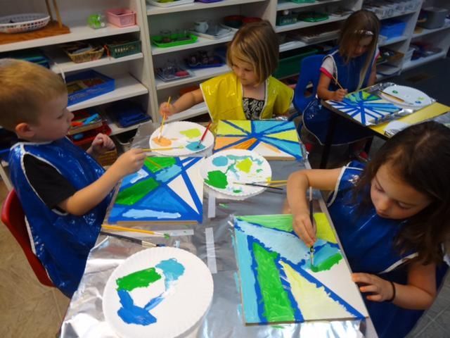 Children painting geometric designs with blue, green, and yellow paint at a table in a classroom