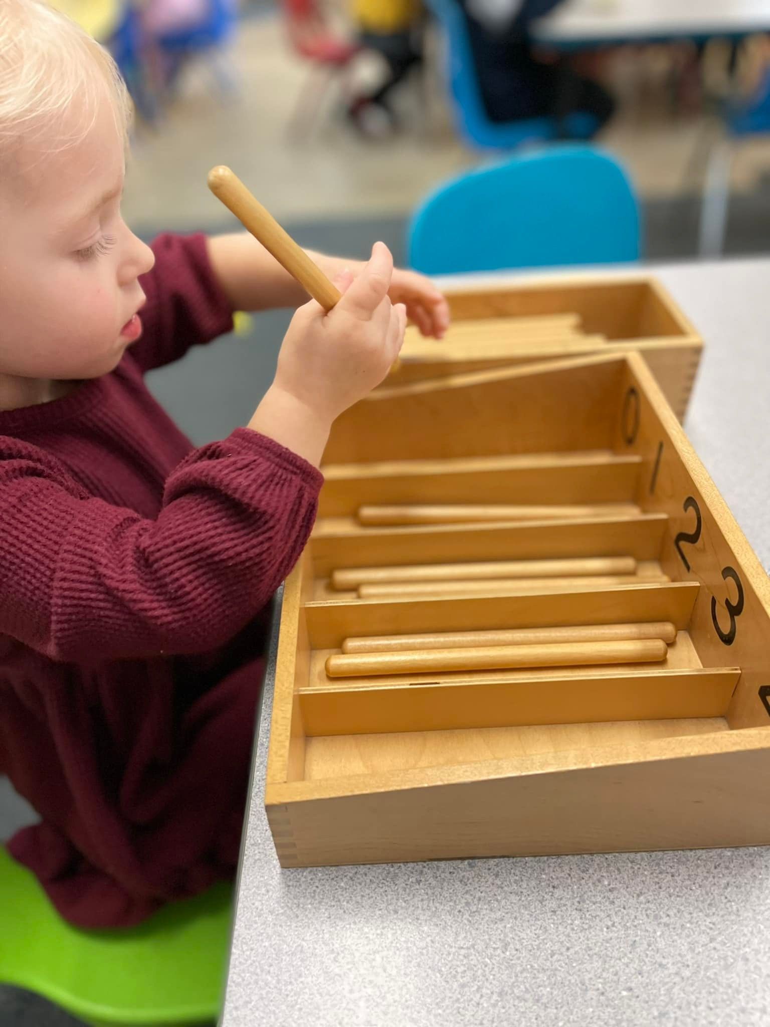 Child in maroon dress playing with wooden rods and box at a table