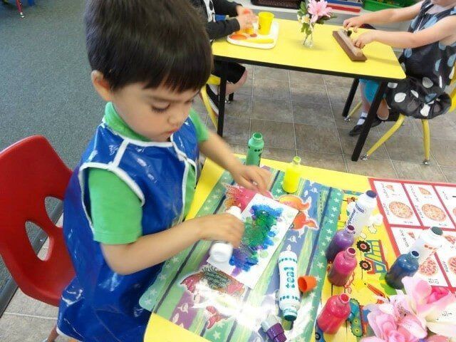 Young boy in blue smock painting at a table in a classroom.
