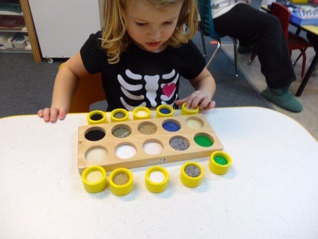 Young girl in skeleton shirt playing with a wooden tray of textured materials in a classroom