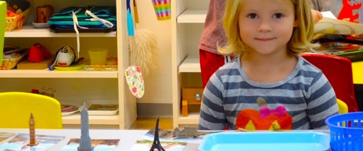 A young blonde girl in a striped shirt smiles at the camera at a table with art supplies