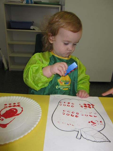 Child in green art smock stamps red paint onto a pear outline and plate