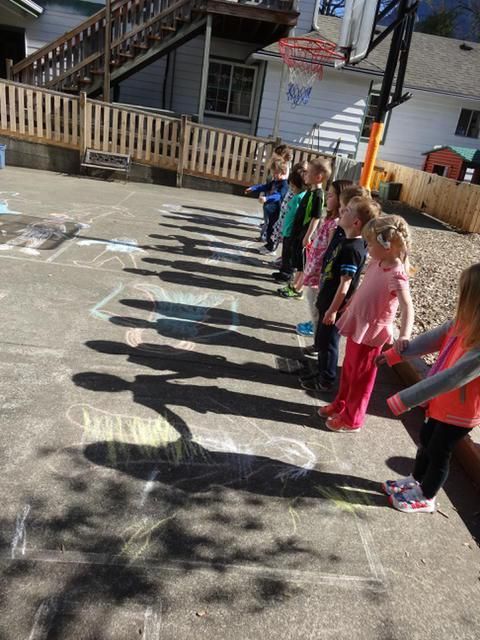 Children standing in a line, tracing their shadows with chalk on a sunny playground
