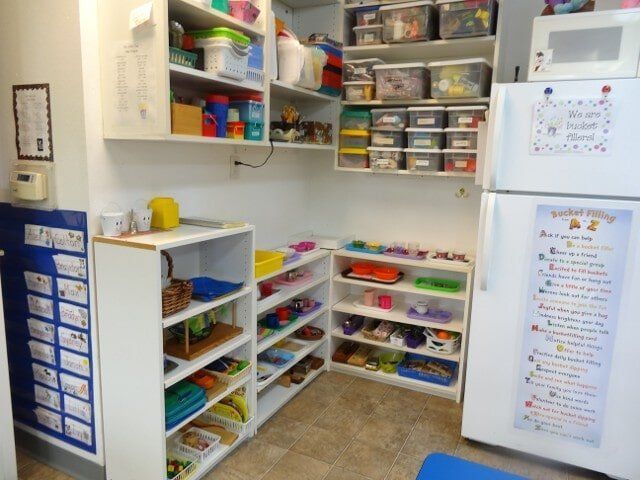 A child's classroom corner with shelves, bins, and a fridge containing toys and supplies