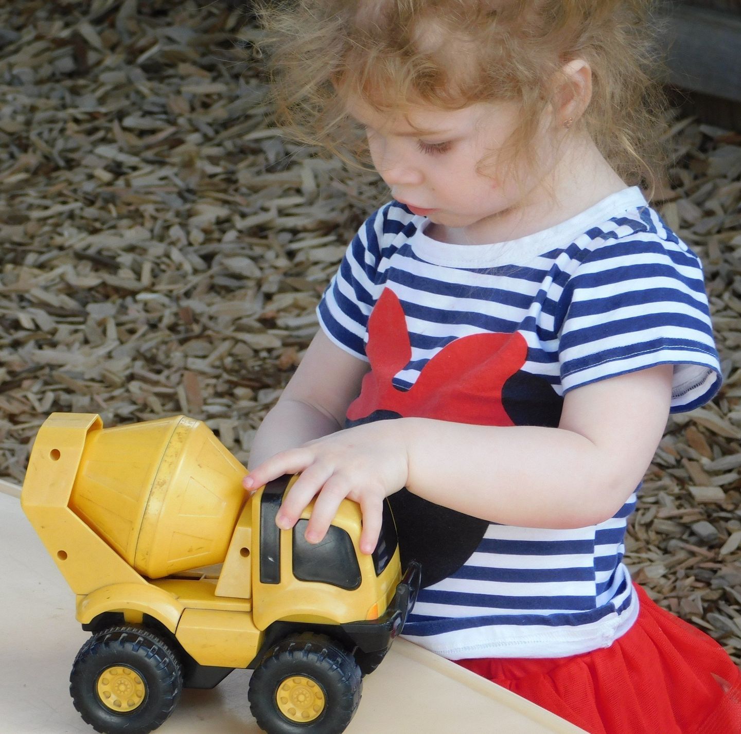 Young child in blue-striped shirt and red skirt plays with a yellow toy cement truck