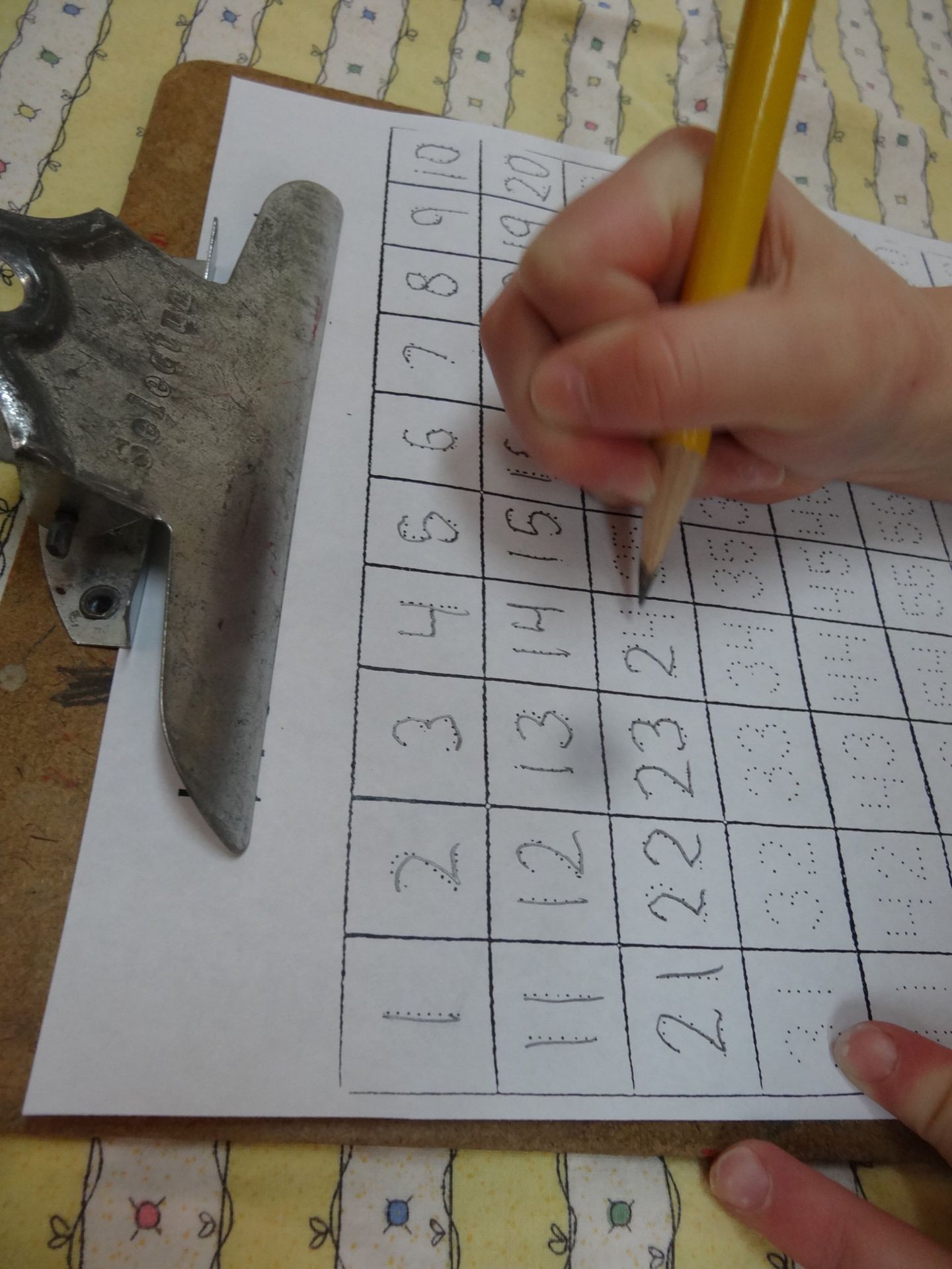 Person's hand holding a pencil, writing numbers on a grid, paper clipped to a clipboard