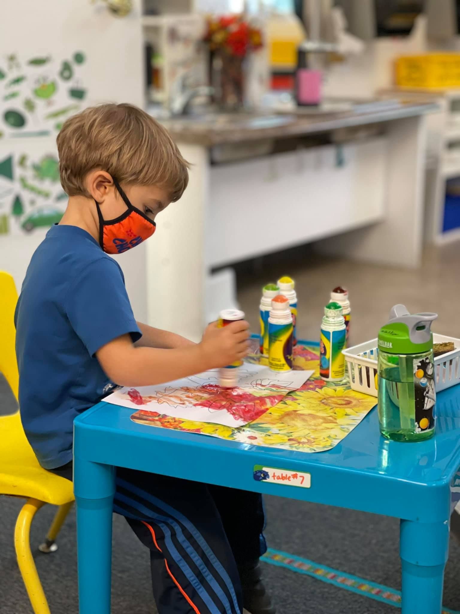 Boy in mask paints at a blue table in a classroom