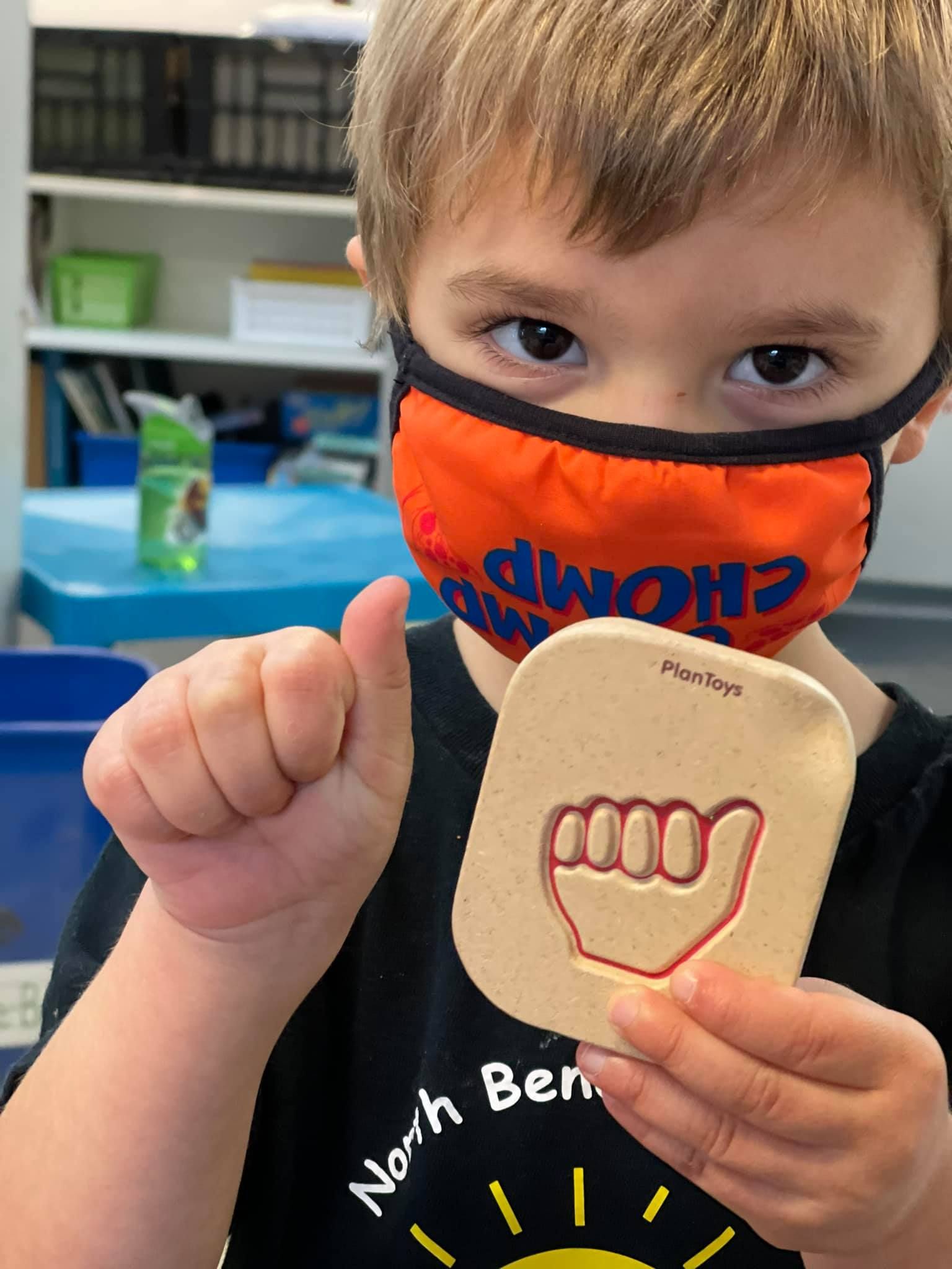 Boy in orange mask gives thumbs up and holds a sign with a hand signing