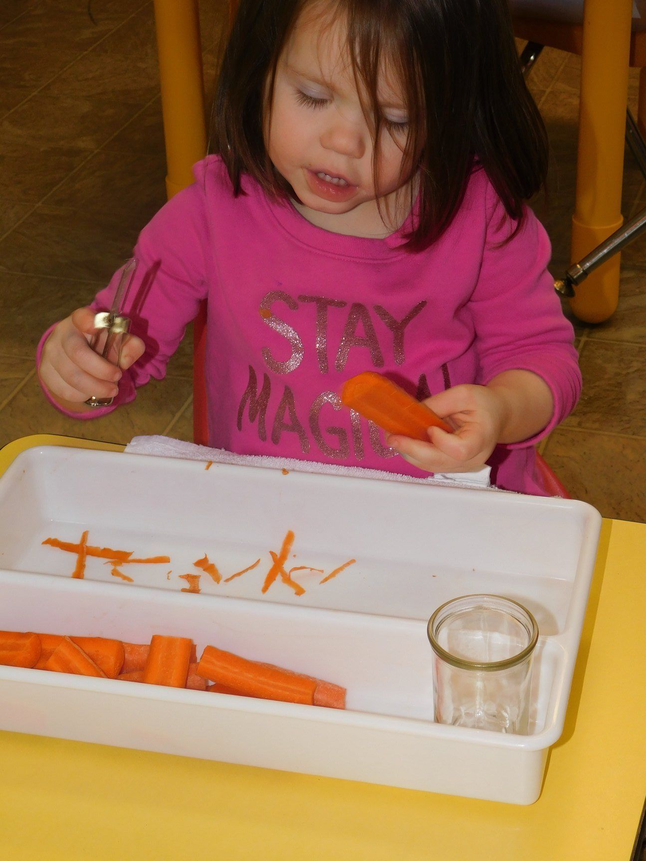 A young girl in pink shirt carefully cuts a carrot with a small knife at a table
