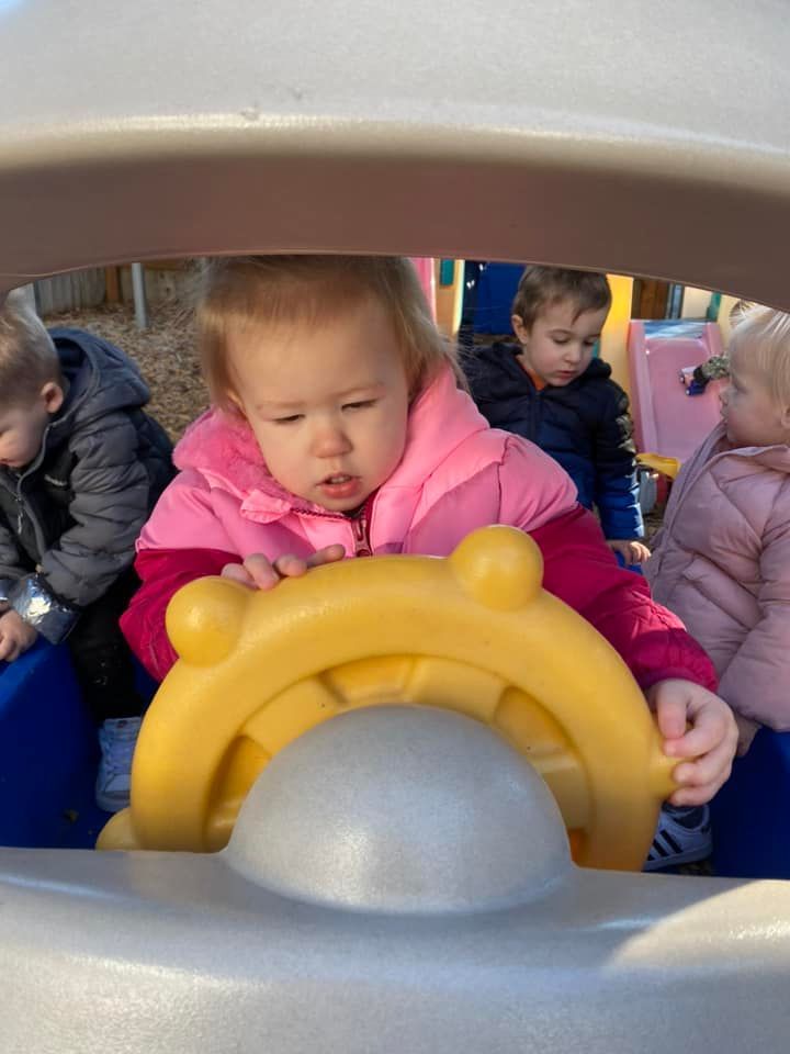 A toddler in a pink vest plays with a yellow steering wheel on a playground