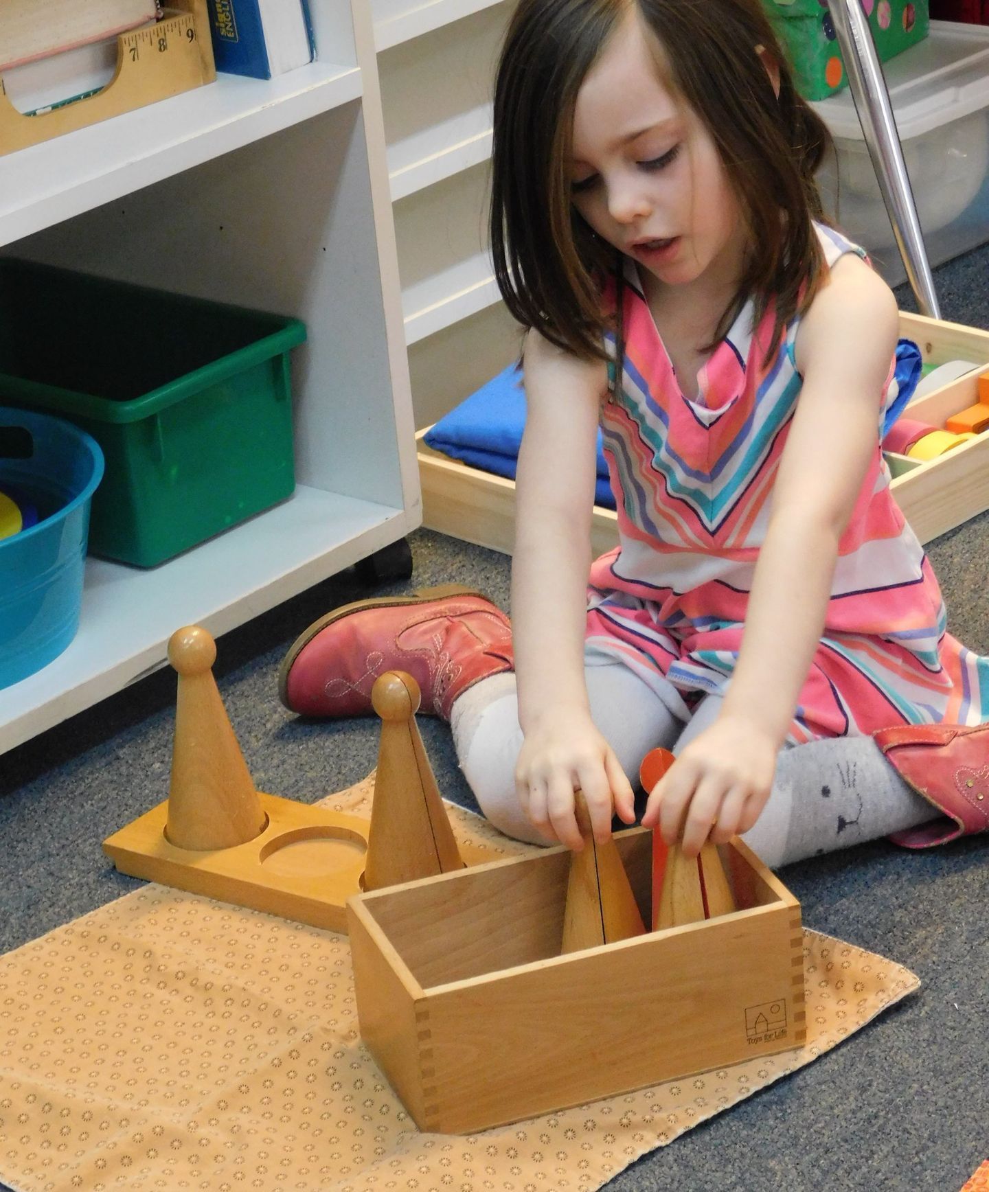 Young girl in a classroom setting, playing with wooden cones and boxes, seated on a mat