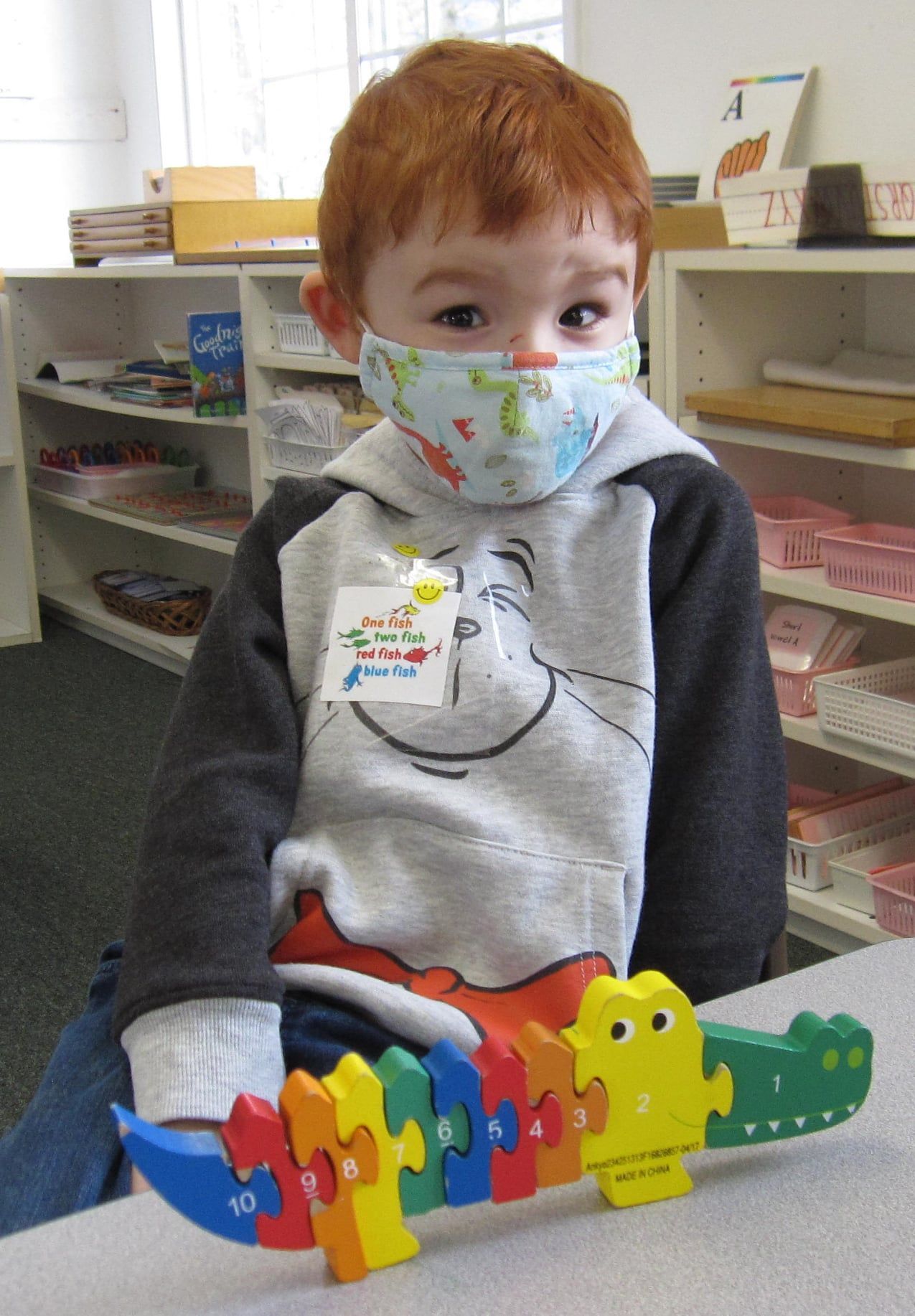 Young child with red hair wearing a mask, holding a colorful alligator puzzle in a classroom