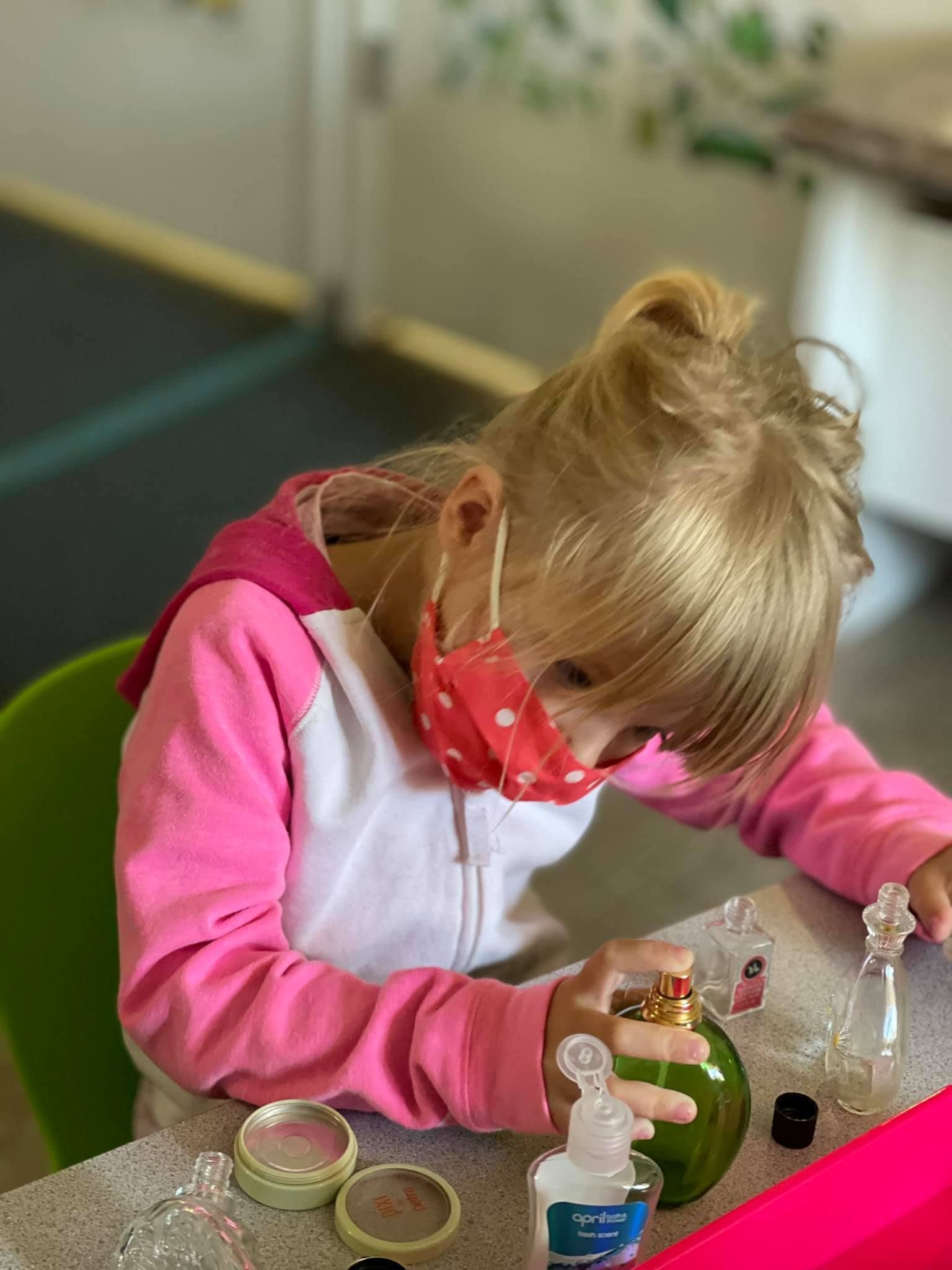 Girl in pink hoodie and mask plays with makeup at a table