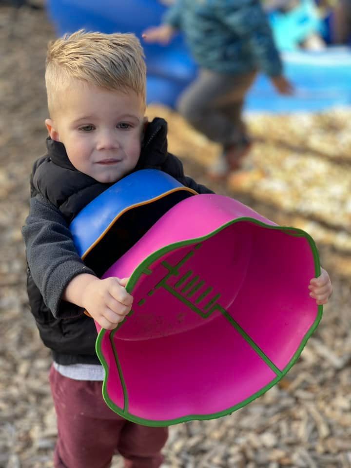 Blond toddler holding large pink and blue toy, looking at camera, outdoors