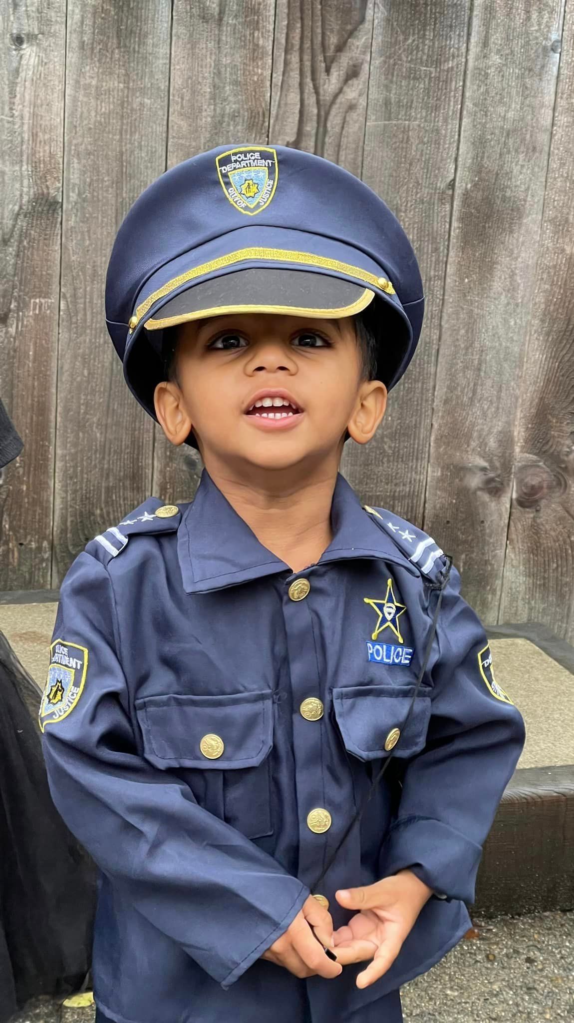 Young child in a blue police uniform and hat, smiling, in front of a wooden wall