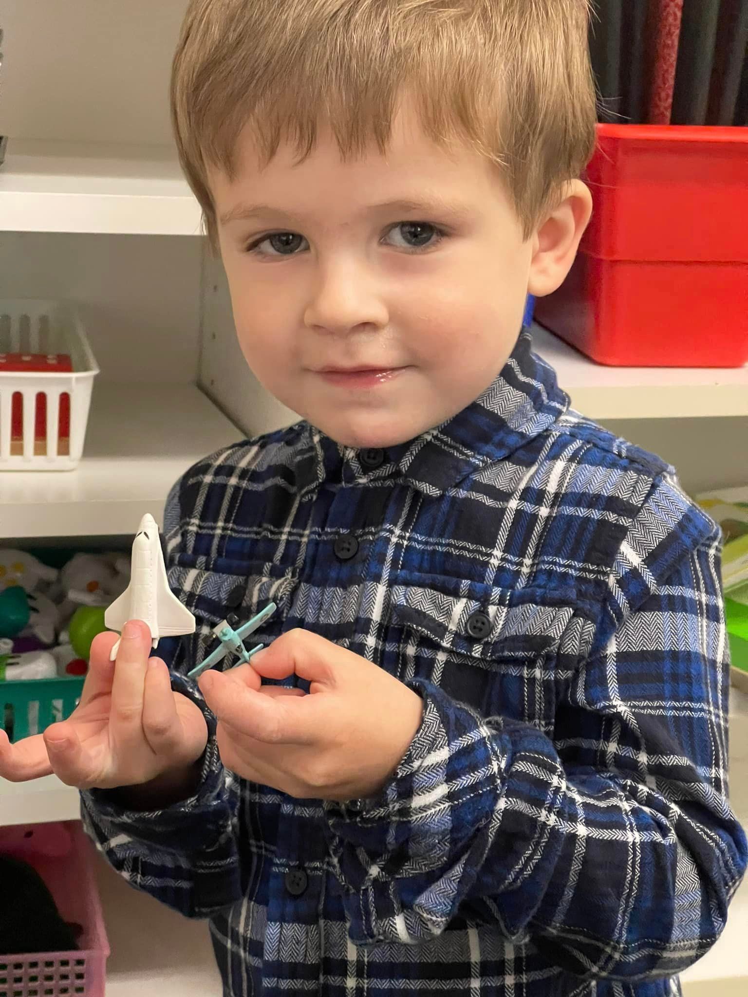 Young boy with short brown hair in plaid shirt, holding a small blue object, smiling at the camera