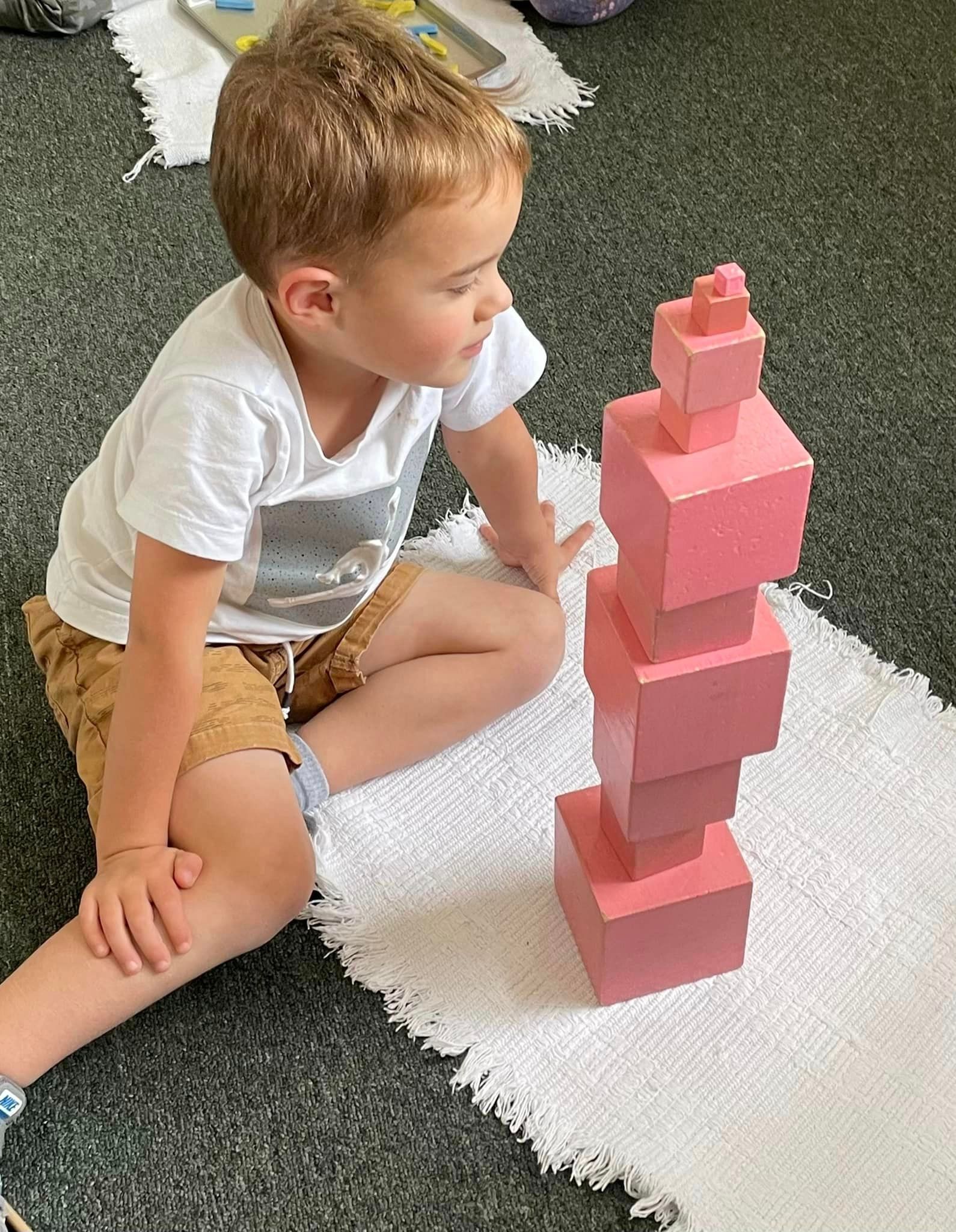Boy looking at a tower of pink blocks while sitting on a white rug