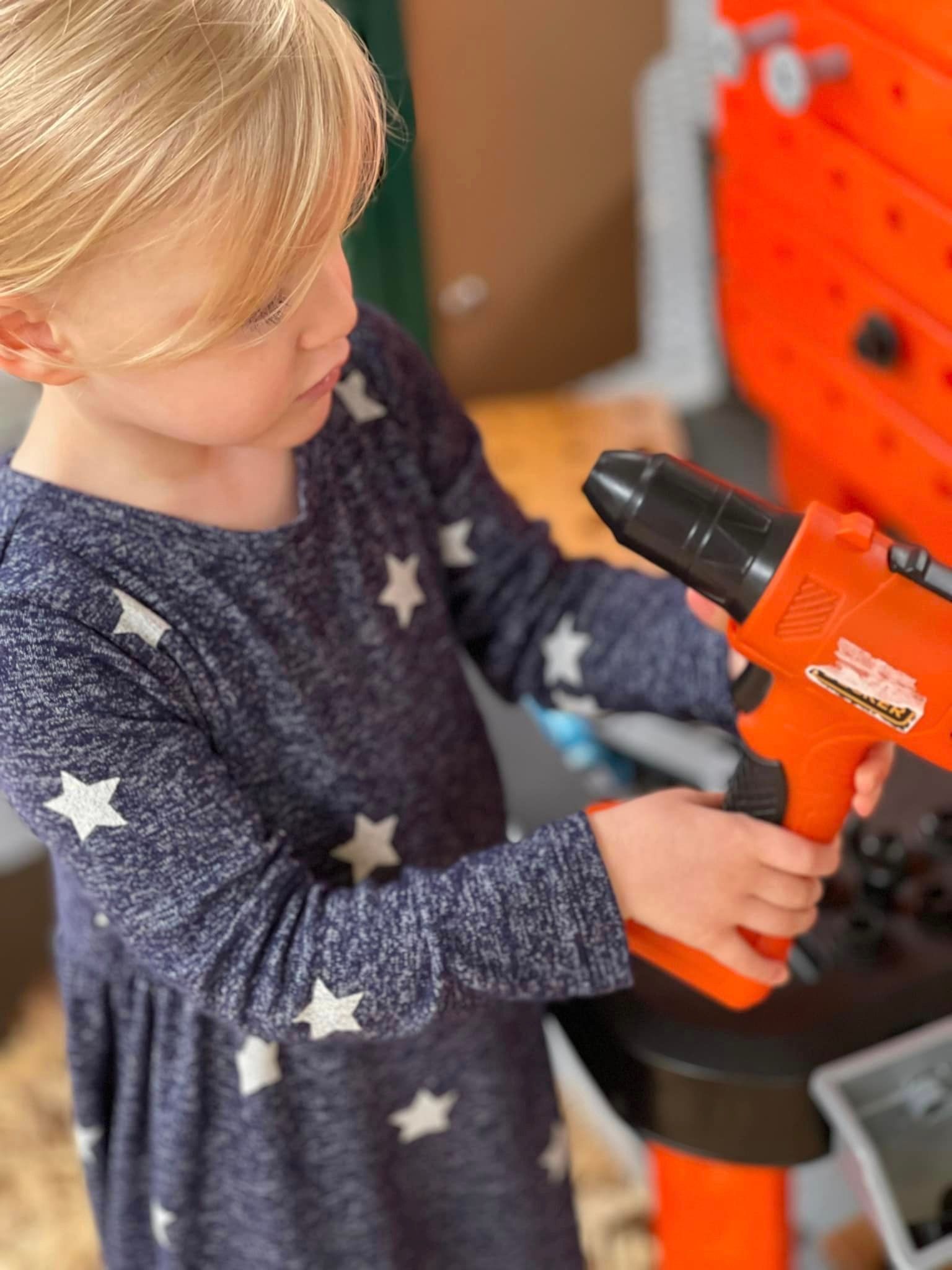 Young girl in star-pattern dress holding an orange toy drill, standing by a tool bench