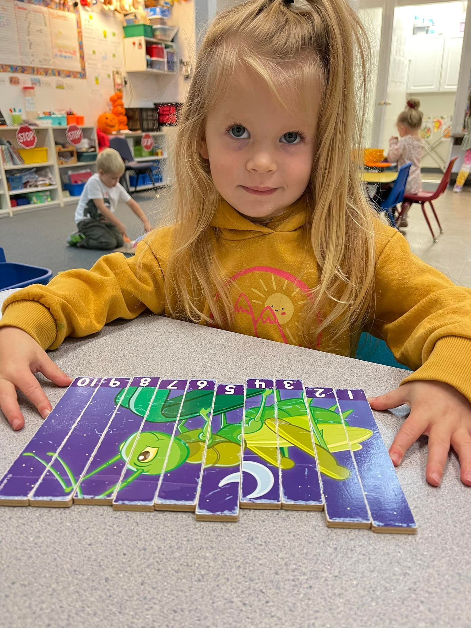 Blond girl smiling at the camera, assembling a caterpillar puzzle on a table at a preschool