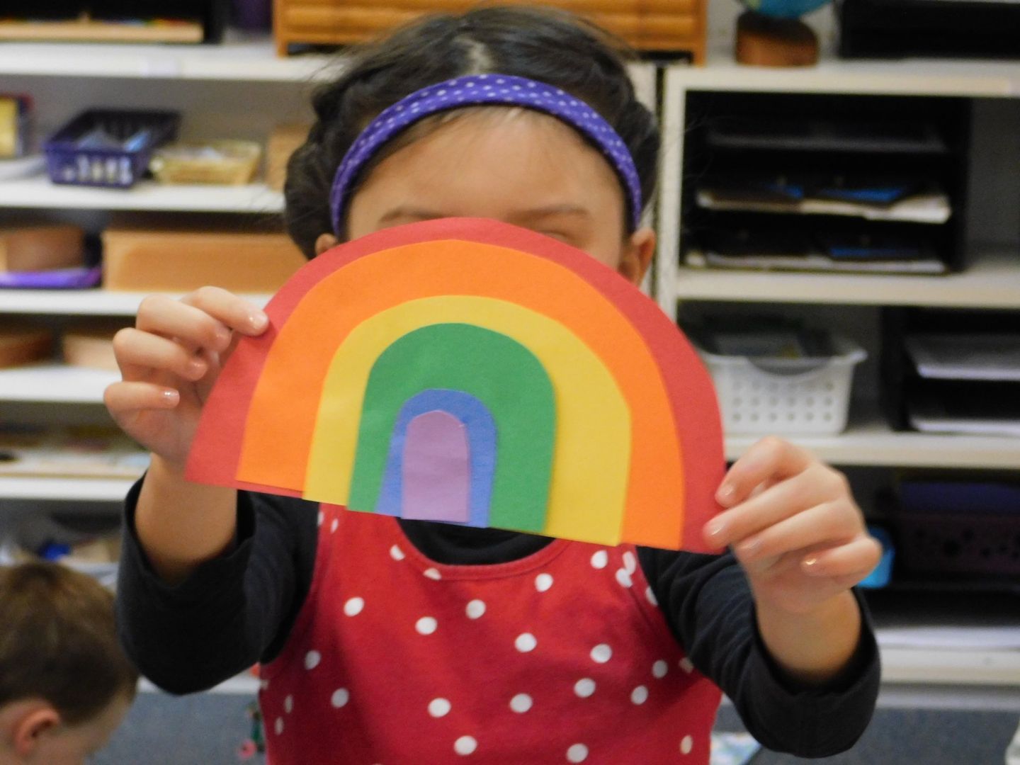 Girl in red polka-dot apron holds up a paper rainbow