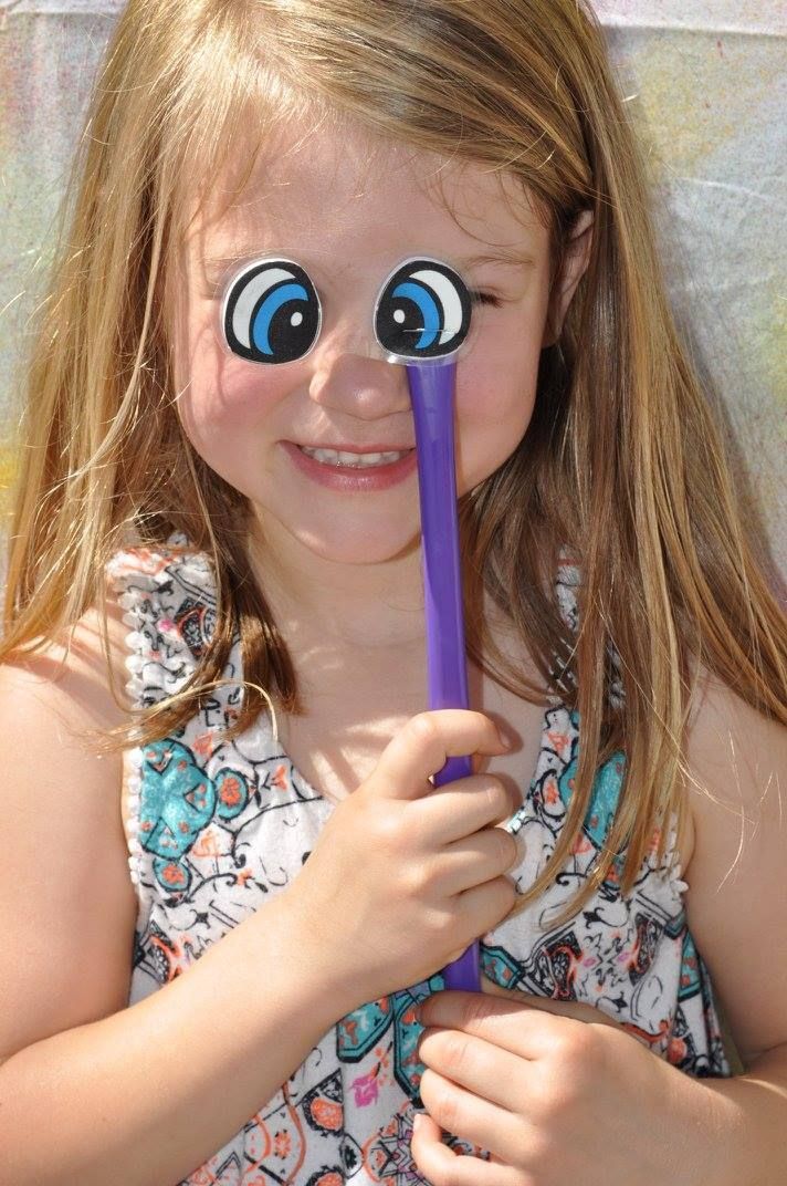 Girl smiling, holding a stick with cartoon eyes over her own, light dress, outdoors