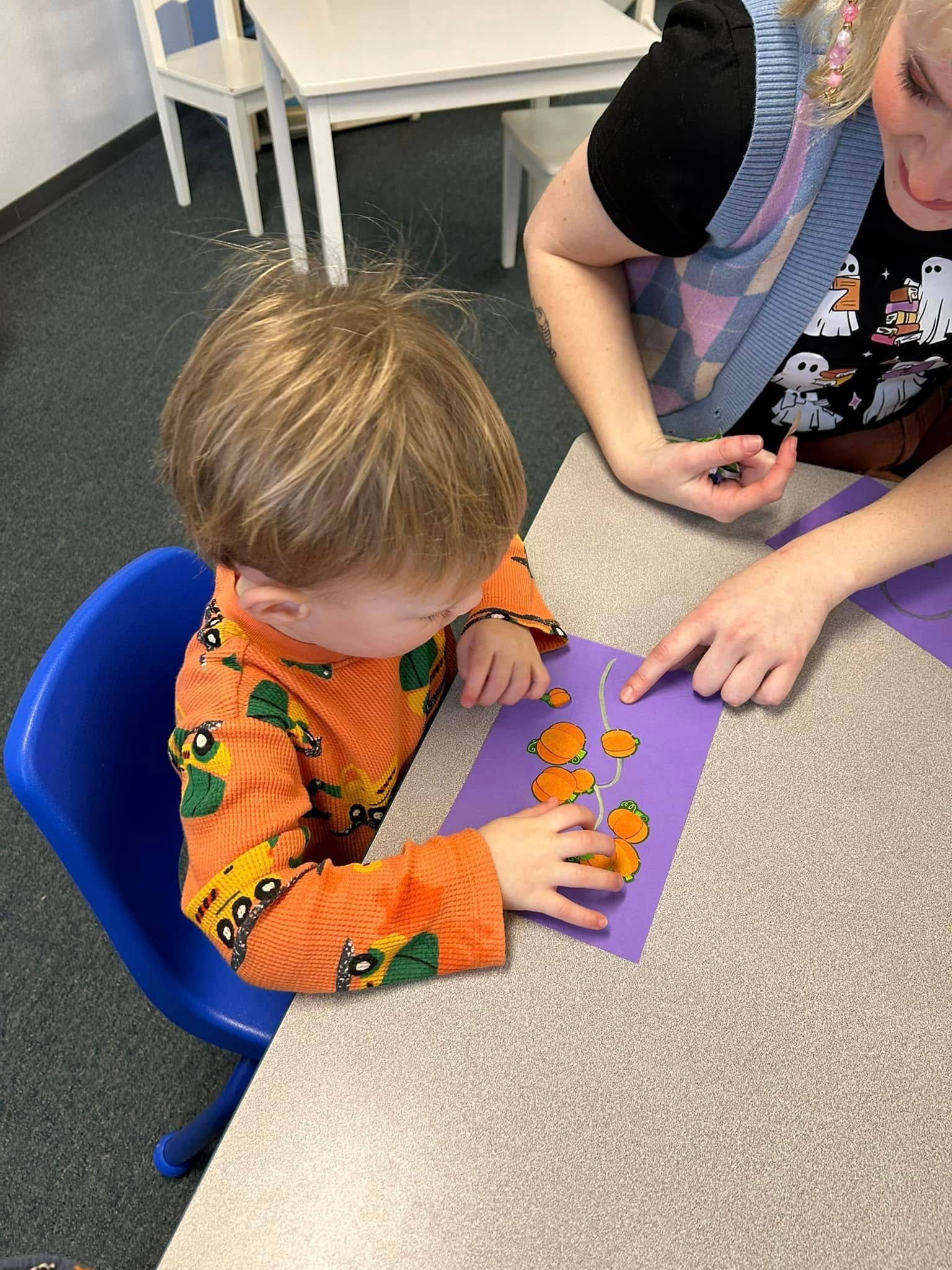 Child in orange pumpkin shirt pointing at a purple card, as an adult points with them