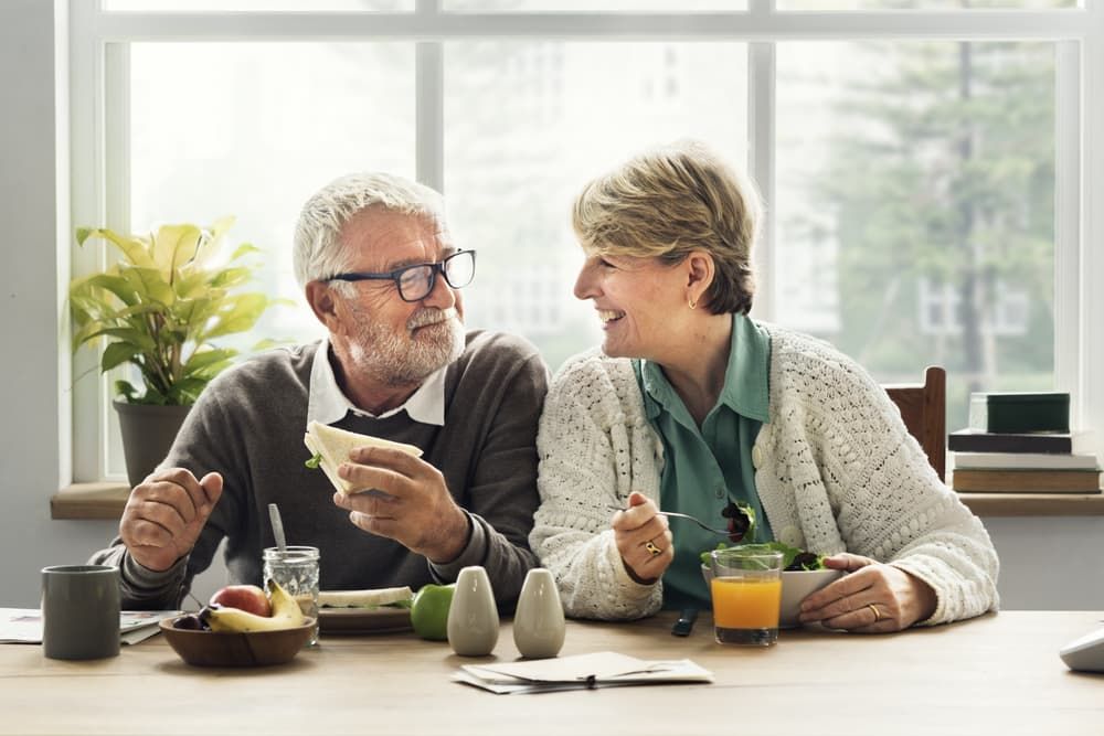 An Elderly Couple Is Sitting at A Table Eating Food - Healthy Cleaning Solutions in Scotts Head, NSW