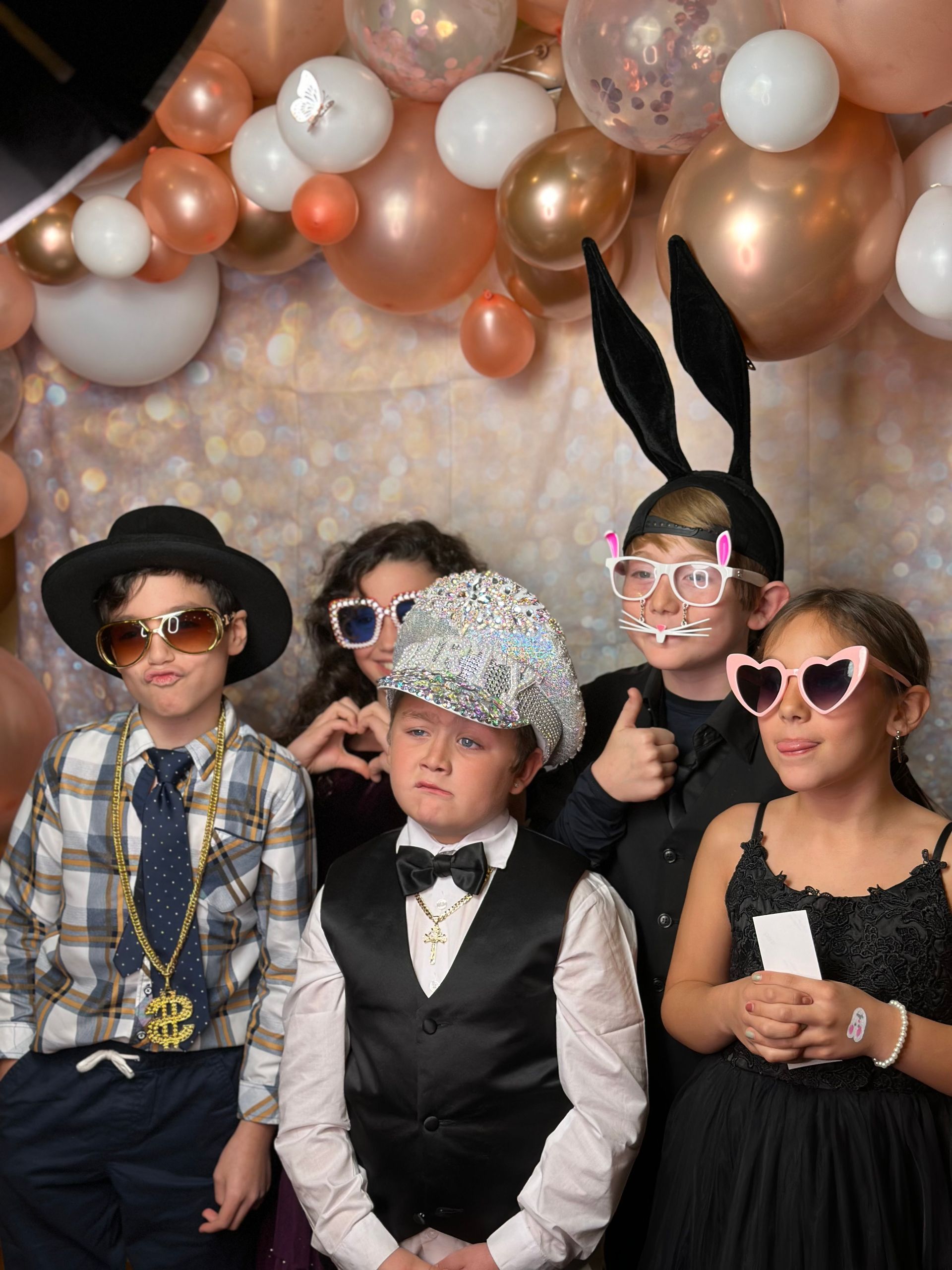 Five children pose for a photo with party props; rose gold and white balloons in the background.