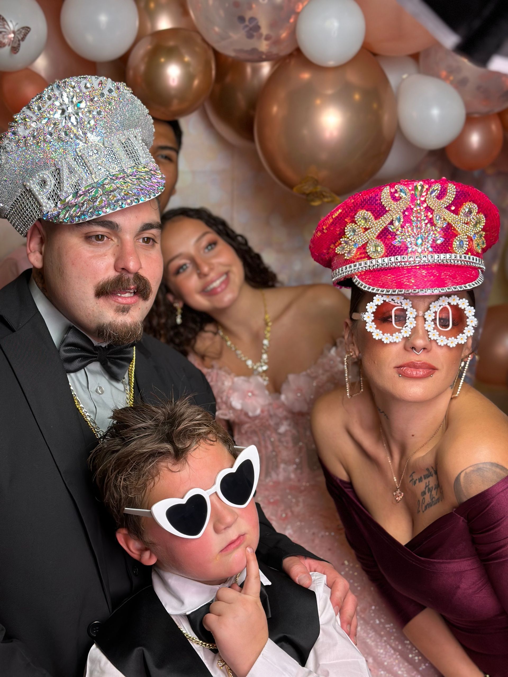 Group photo booth shot with people in party hats, sunglasses, and formal wear, smiling and posing. Balloons in the background.