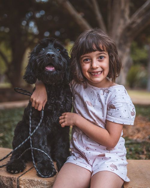 A little girl is sitting next to a black dog on a leash.
