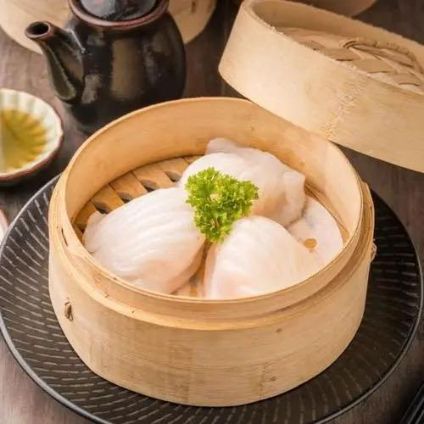 Steamed dumplings in a bamboo basket with a sprig of parsley on a tray, beside a teapot.