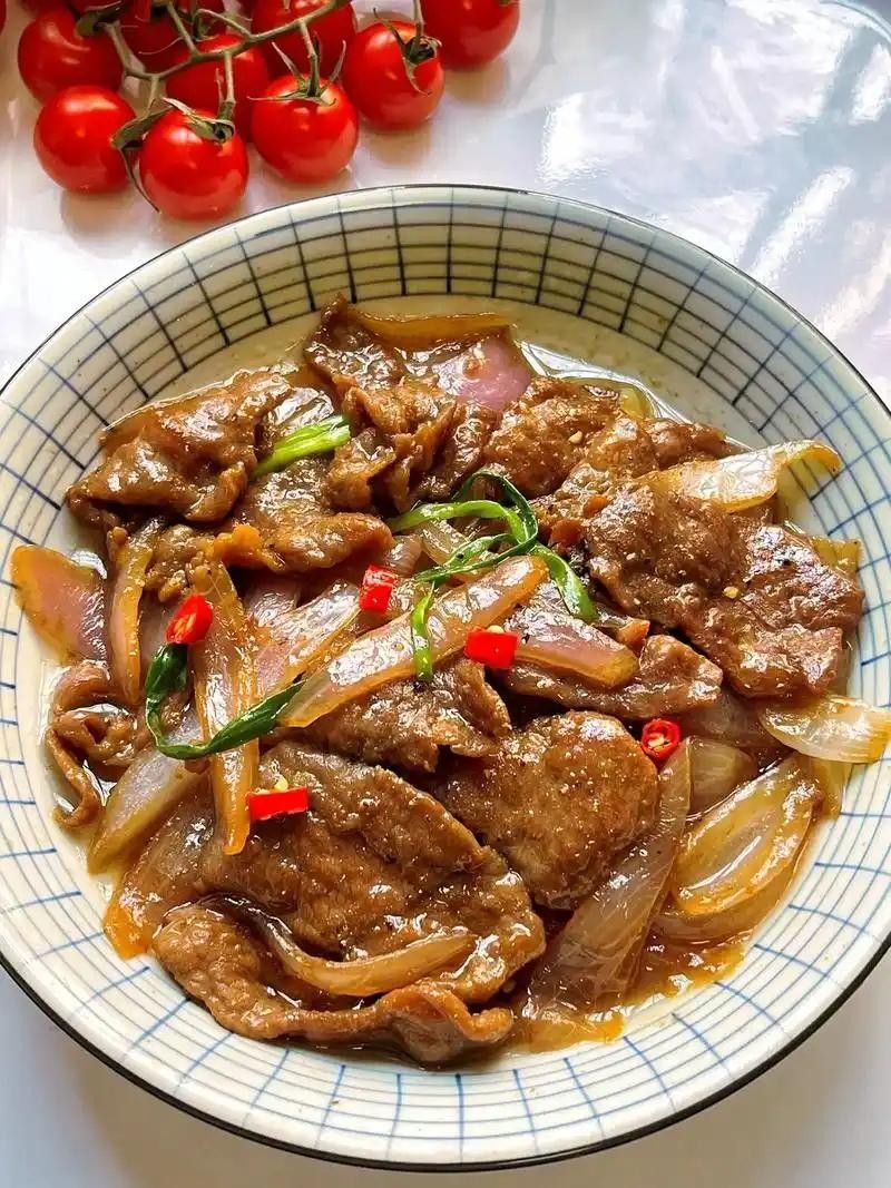Stir-fried beef with onions and chili peppers in a bowl, with a bunch of cherry tomatoes nearby on a white surface.