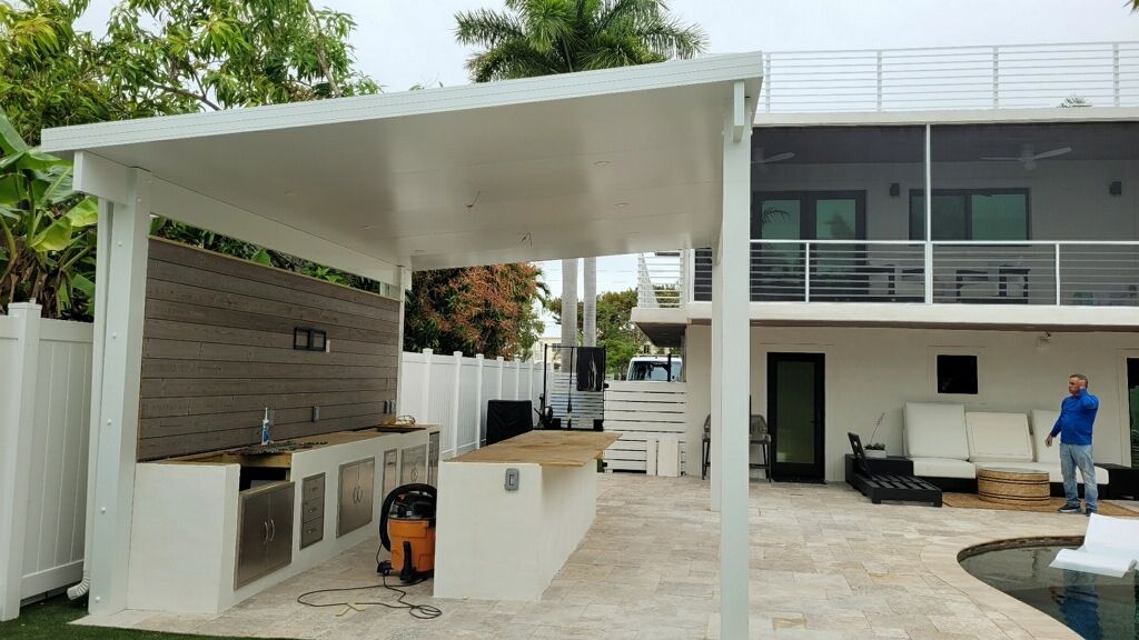 A white covered outdoor kitchen and bar area featuring a wood-paneled backsplash, located next to a swimming pool.