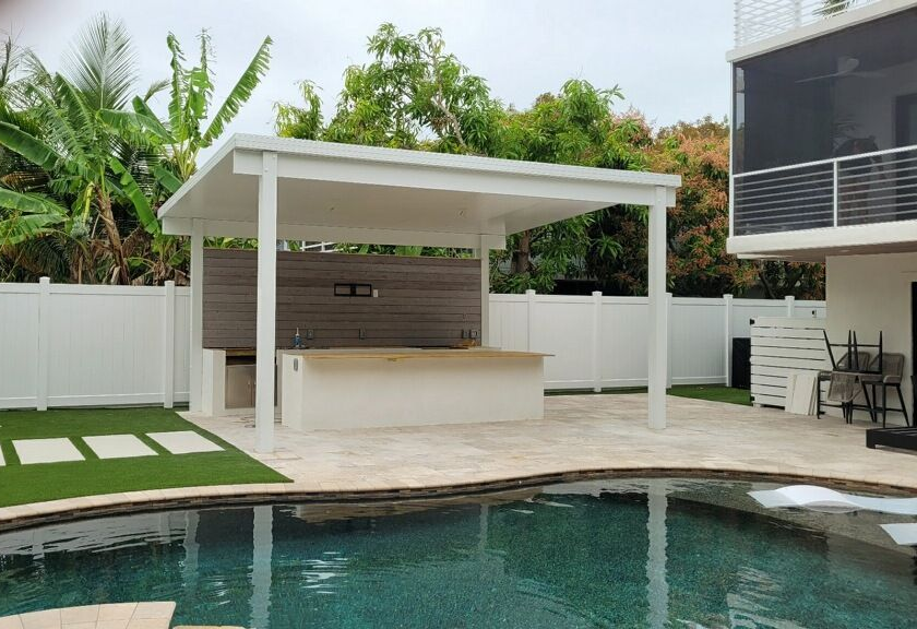 An outdoor swimming pool with a covered bar and seating area, surrounded by a white fence and lush green plants.