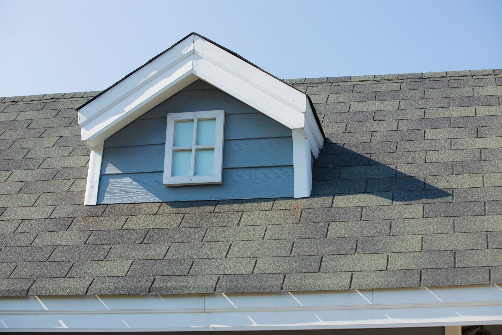 Blue dormer with white trim and window on gray shingled roof, against a clear sky.