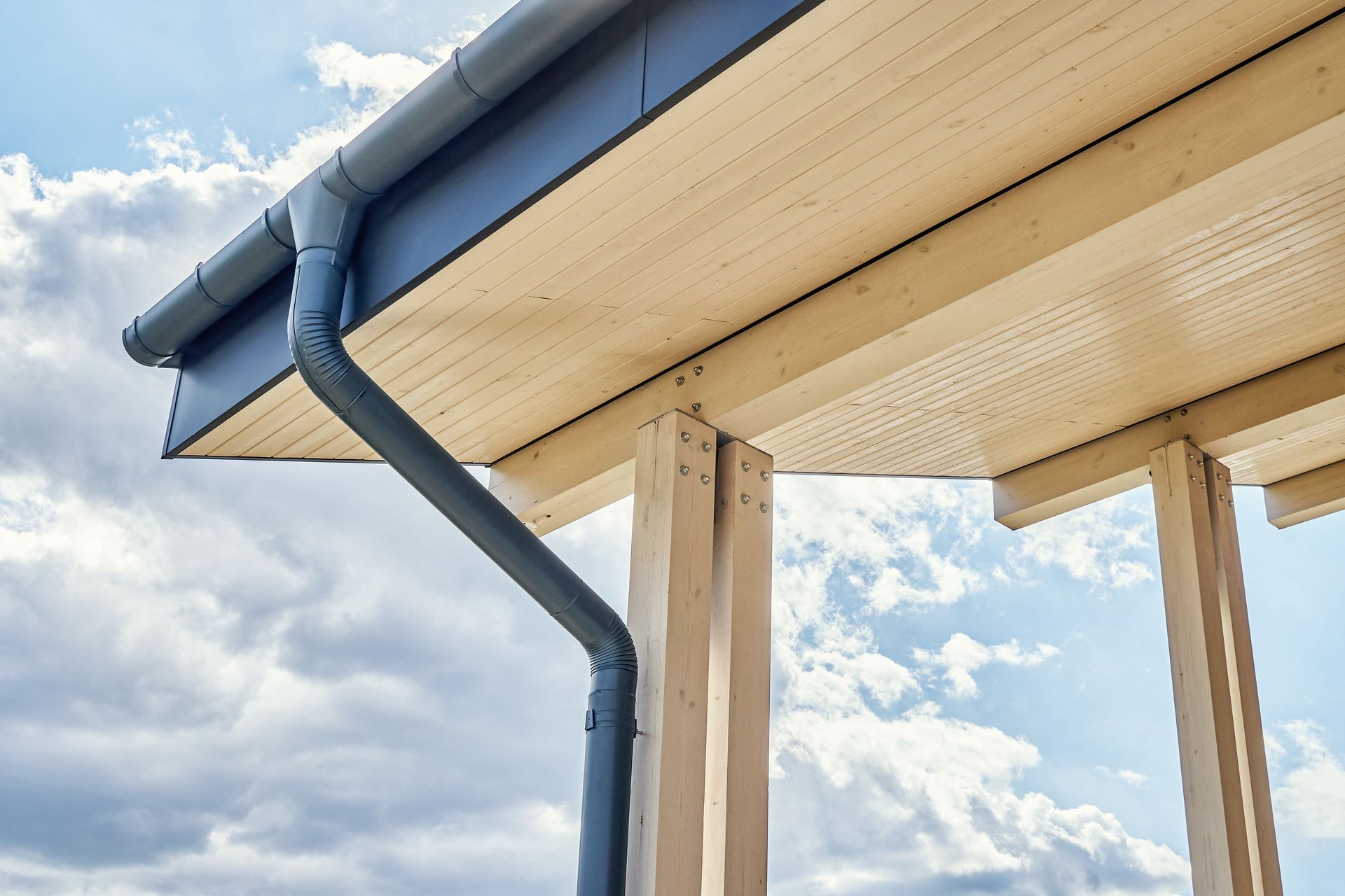 Gray rain gutter and downspout on a light-colored wooden porch roof against a cloudy sky.