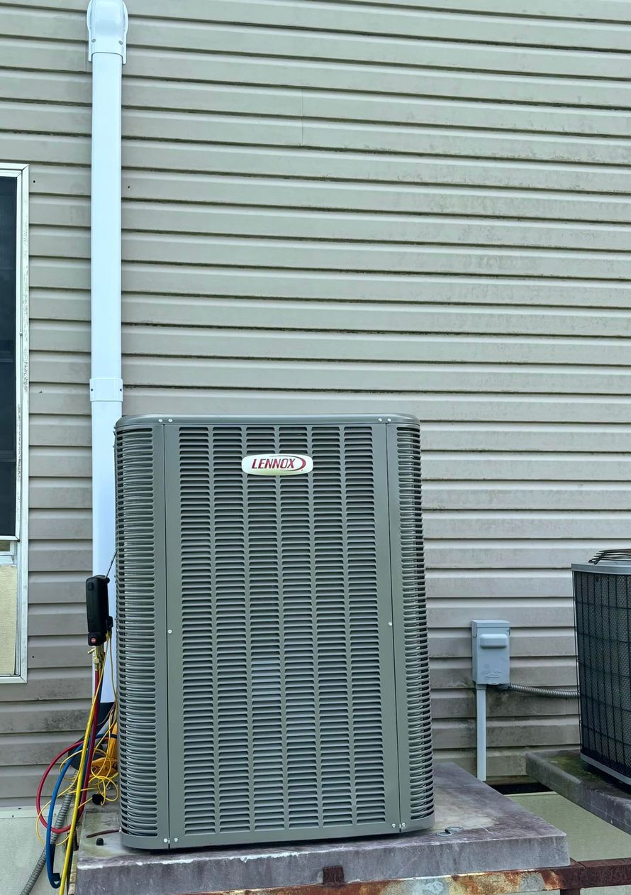 Air conditioning unit outside a building with light gray siding and a white pipe.